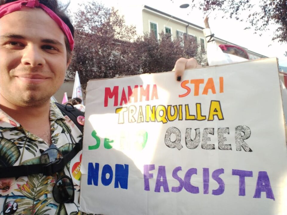 Giovanni, un demiguy. tiene un cartello con su scritto "Mamma, stai tranquilla. Sono queer, non fascista".